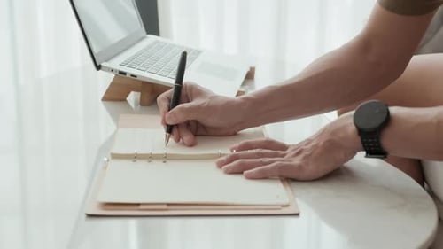 Adult Writes in Notebook With Laptop Computer