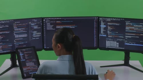 Asiatische Frau arbeitet mit Tablet, während sie Code mit mehreren Computerbildschirmen auf grünem Bildschirm schreibt