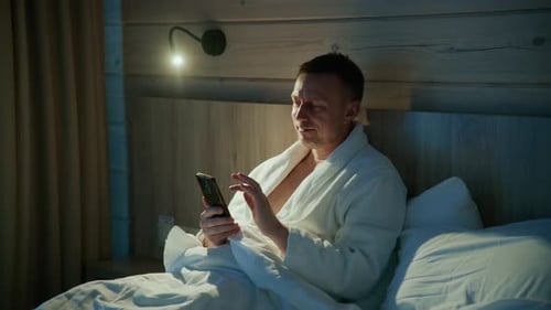 A Man Wearing a Cozy Bathrobe is Comfortably Using His Smartphone While Relaxing in Bed During the