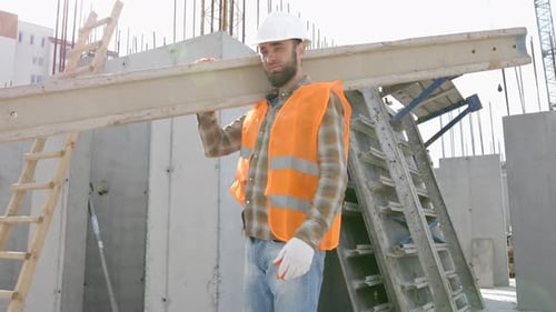 Builder man in hardhat and vest carrying timber on building site in his working day