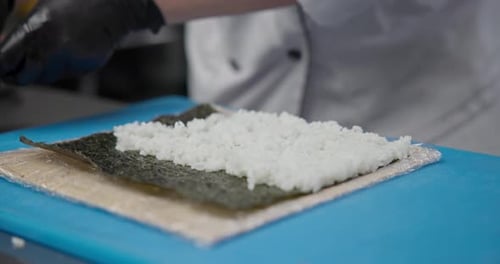 Un chef portant des gants en nitrile noir prépare des sushis dans la cuisine en étalant du riz cuit sur du nori