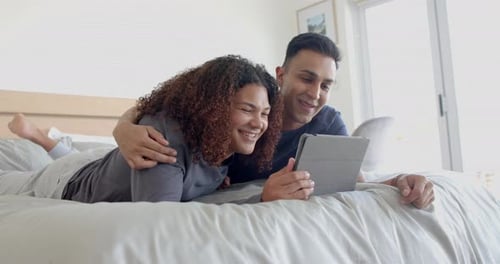 Fröhliches, vielfältiges schwules männliches Paar, das mit Tablet auf dem Bett liegt und sich morgens umarmt, Zeitlupe