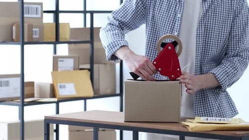 Man using dispenser with adhesive tape to seal cardboard box and sticking barcode onto it