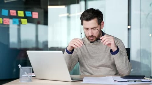 Exhausted businessman working on laptop and paperwork at desk in modern office. Frustrated mature