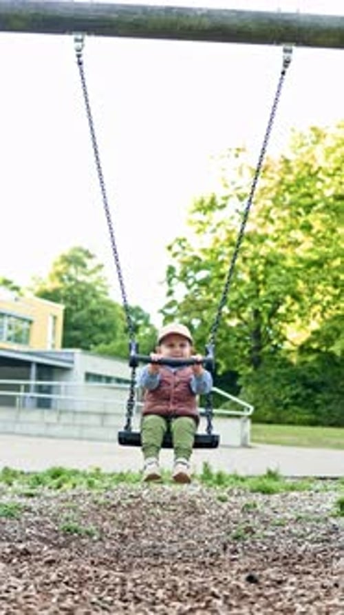 Toddler in blazer sits on swing in city park. Boy swings on swing on playground