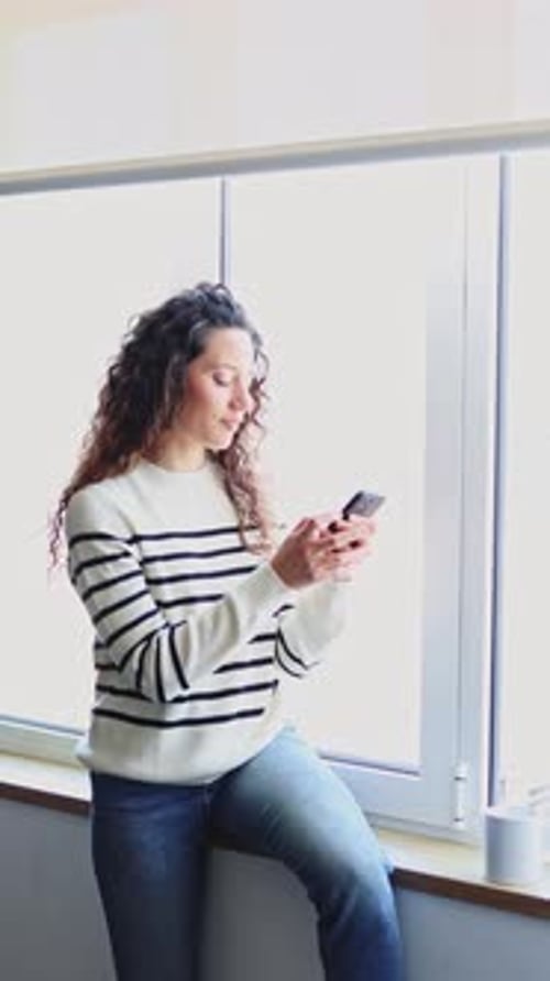 Woman Uses Smartphone and Drinks Near a Window