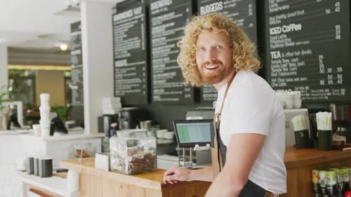 Portrait of happy caucasian male barista, smiling with arms crossed behind counter in cafe