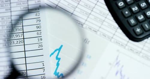 A hand with a magnifying glass passes over a financial newspaper and financial bag with a business