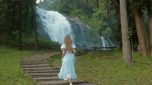Woman in Blue Dress Admiring Waterfall in Lush Green Forest Serene