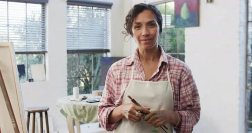 Portrait of biracial female artist in apron holding brushes in studio, slow motion