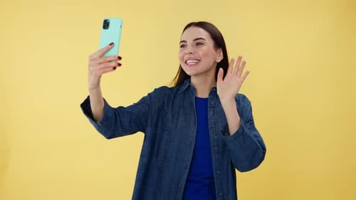 Dama feliz agitando la mano durante una videollamada en un teléfono celular moderno en el estudio