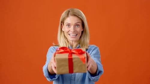 Smiling Woman Presents Gift on Orange Background
