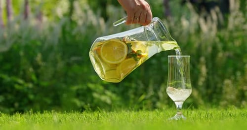 Hand Pours Refreshing Lemonmint Lemonade Into Glasses on a Vibrant Green Lawn in a Sunny Outdoor