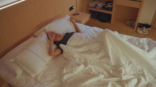 Caucasian Woman Sprawled on Hotel Bed Arms Out Under Half Pulled Duvet Warm Wood Headboard Suitcase