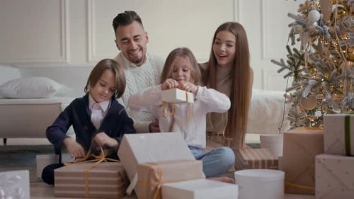 Young Parents And Two Children Sit On Floor And Unpack Boxes Of Gifts
