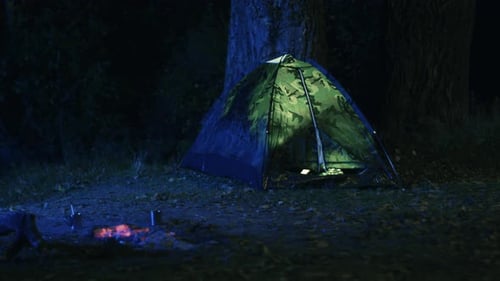 Light is turned off in a tent next to a burning campfire at night in a forest.