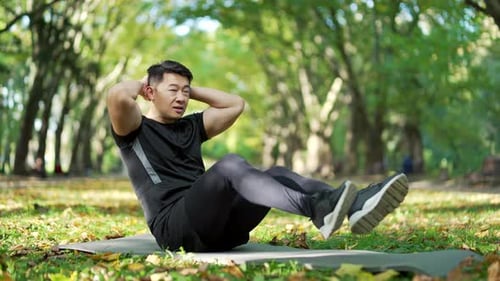 Asian man exercises abdominal muscles while sitting on a mat in an urban city park. Handsome male