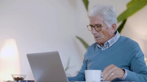 Adult with Gray Hair Drinks Coffee While Using Laptop