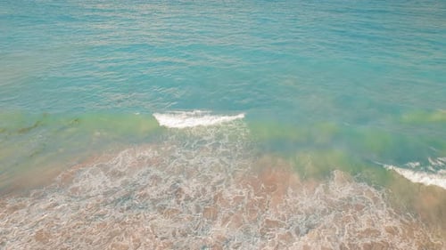 Top down view of turquoise ocean waves rolling onto sandy shoreline in sunlight