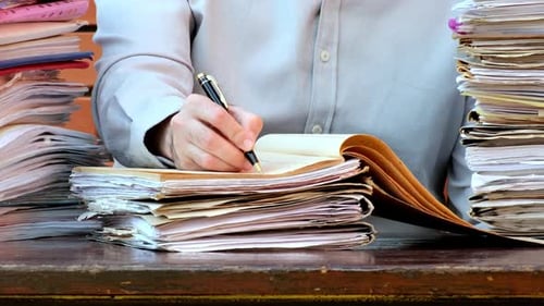 Writing in Notebook Surrounded by Stacks of Paper