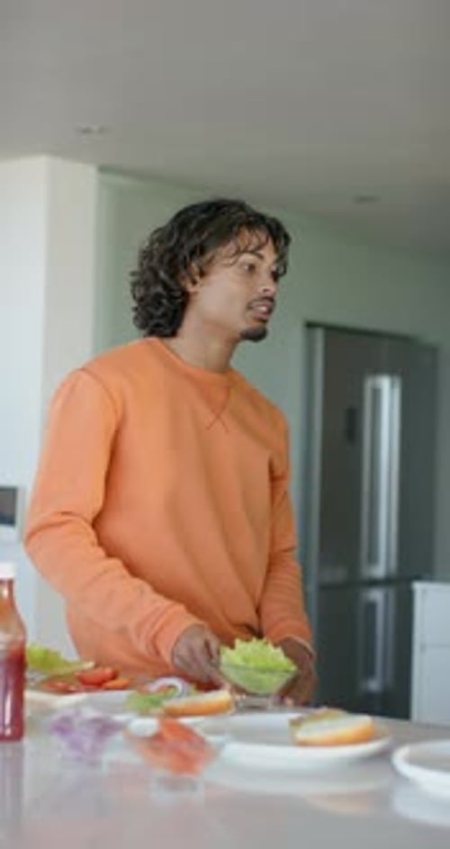 Vídeo vertical: Homem adulto com camisa laranja olhando para a ilha da cozinha estendendo uma tigela de alface servindo amigo