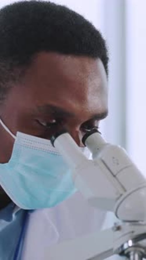 African American Young Biologist Examining Vaccine Test with Microscope Wearing Face Mask and Gloves