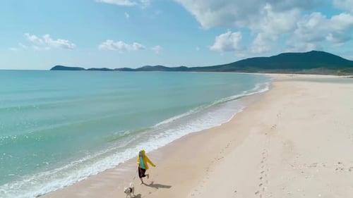 Aerial View of a Young Woman in Yellow Jacket Running on Beach with Her Dog