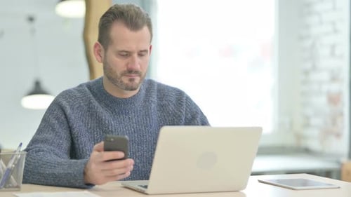 Man Using Mobile Phone at Desk with Laptop