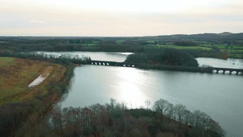 Luftpanorama-Steinbogenbrücke, die sich über einen breiten See in der Nähe der bewaldeten Küste und Sonnenlicht erstreckt