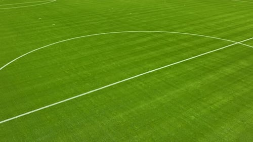 Close-up, low-angle aerial view of a portion of a lush green soccer field. It shows part of the whit