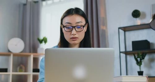 Portrait of concentrated skilled young asian woman in glasses working on laptop in contemporary home