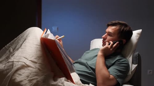 Man Talking on Phone While Reading Documents in Bed