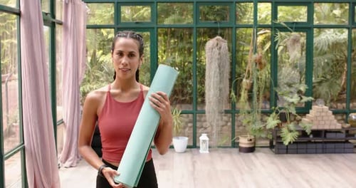 Smiling woman holding yoga mat, preparing for yoga session in glass house, copy space