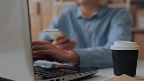 An Employee Manager Works At A Laptop Computer In A Warehouse Against The Background Of Cardboard