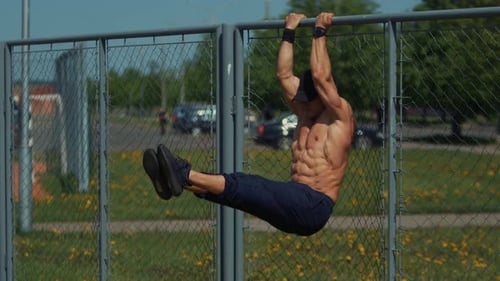 Young Muscular Man is Training on Outdoor Sports Field and Doing Hanging Leg Raises While on Fence