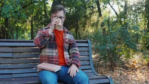 Young Man with Down Syndrome in Checkered Shirt Drinking Coffee Outdoors in the Public Park