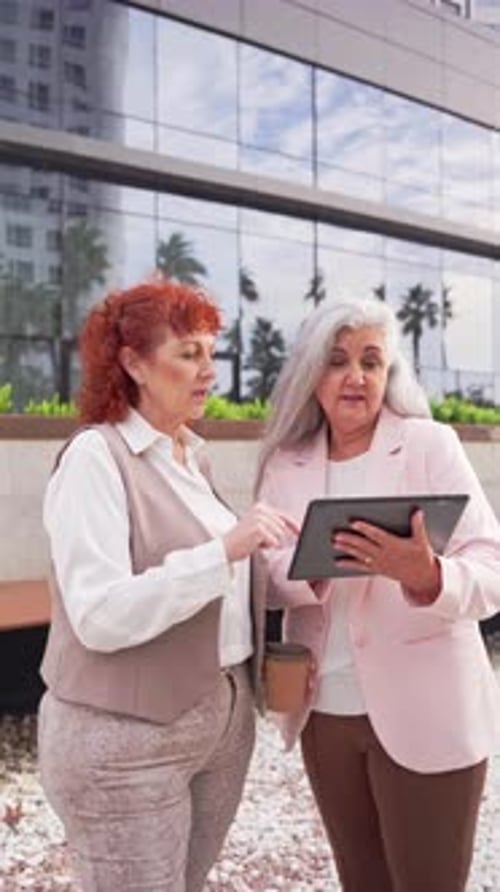 Two Professional mature women standing outside office collaborating and reviewing data on tablet