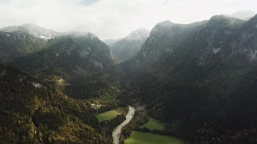 The Drone Flies Over a Narrow Mountain River Between Rocky Alpine Mountains Covered with Snow and