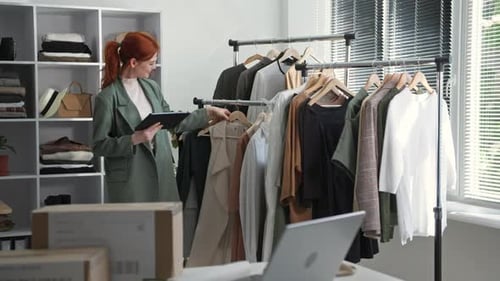 Jovem proprietária de uma pequena empresa usando óculos verifica cabides com tablet na mão e roupas