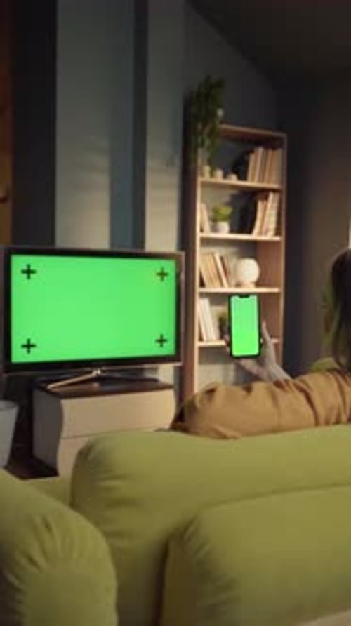 Woman Sitting on a Sofa in a Living Room Holding a Smartphone with a Green Screen While a Television