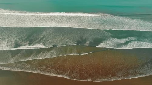 Aerial view of ocean waves rolling to shore on dark sandy tropical beach