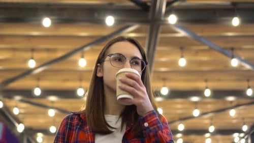A Young Girl Walks Along the Alley of Light Bulbs with Coffee in Her Hands