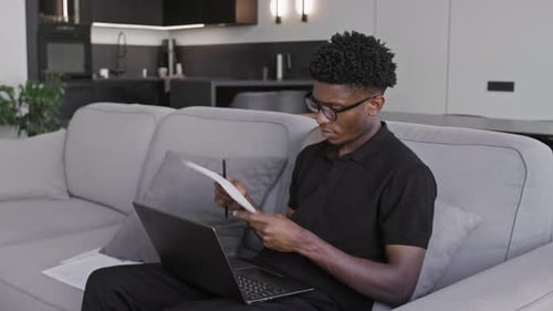 Man Works On Laptop On Couch With Papers