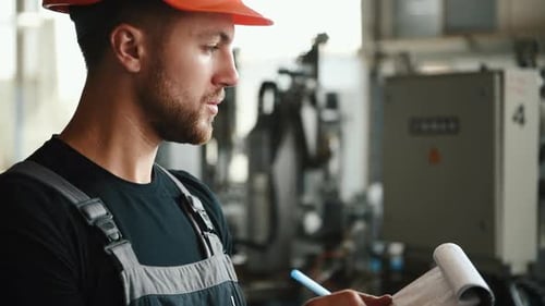 Manager in orange hard hat stands in the factory with machines and writes information in notepad
