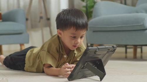Close Up Of Asian Kid Lying On The Floor Using A Tablet At Home