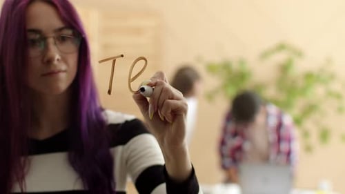 Caucasian Woman with Purple Hair Writes the Word Teamwork with a Marker on the Glass Against
