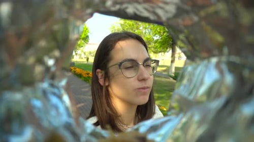 A Young Woman with Glasses Eats Potato Chips From a Pack While Walking in the Park View of a Woman