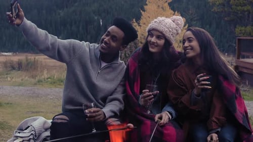 Happy young friends take a selfie with camera phone at fire pit outside cabin