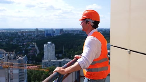 Happy Male Builder Wearing Helmet and Protective Glasses Standing on the Roof Looking at the View