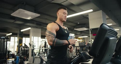 A Young Man with a Tattoo is Jogging on a Treadmill Warm Up in the Gym Cardio